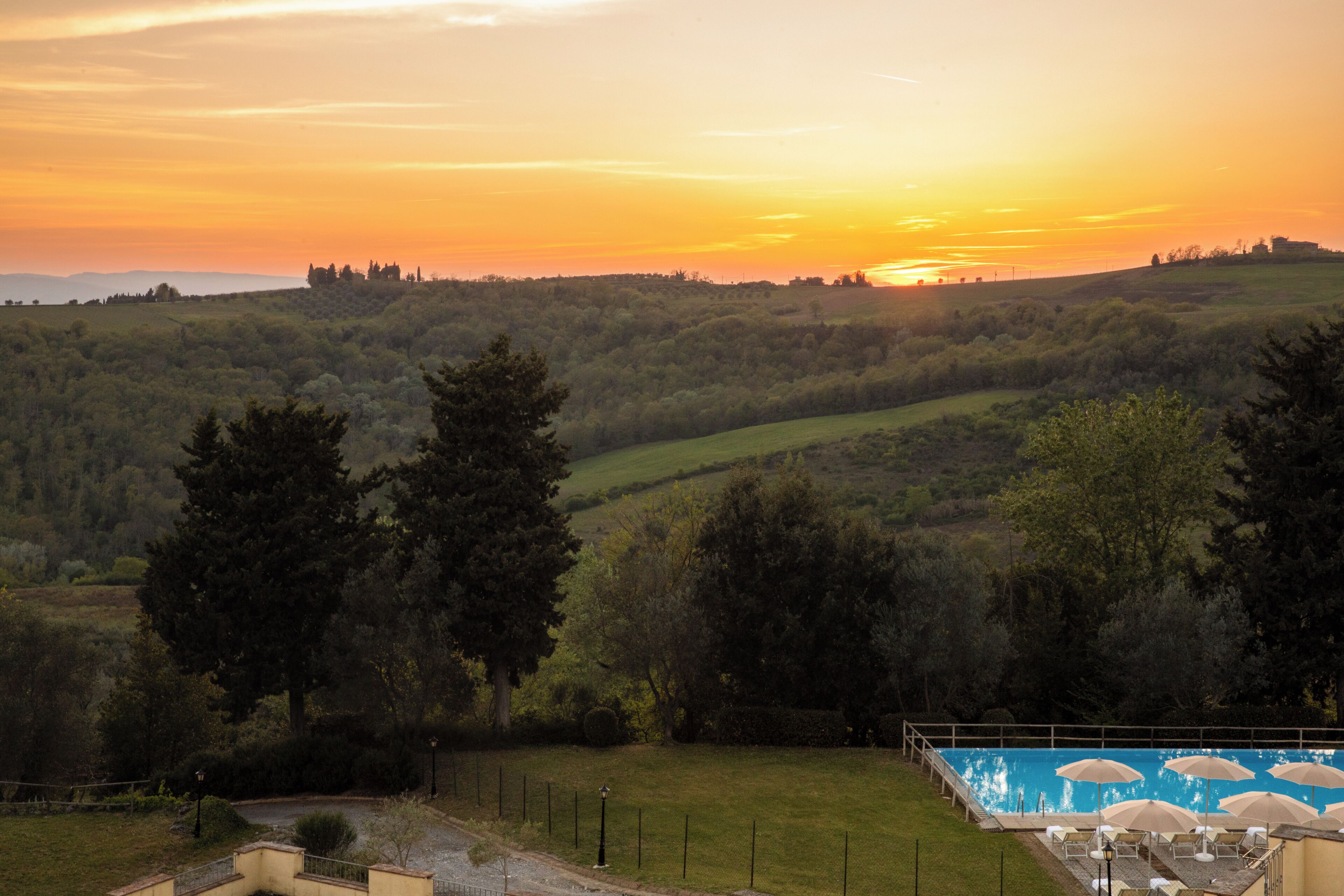 A pool area with umbrellas, surrounded by a hilly landscape and trees.