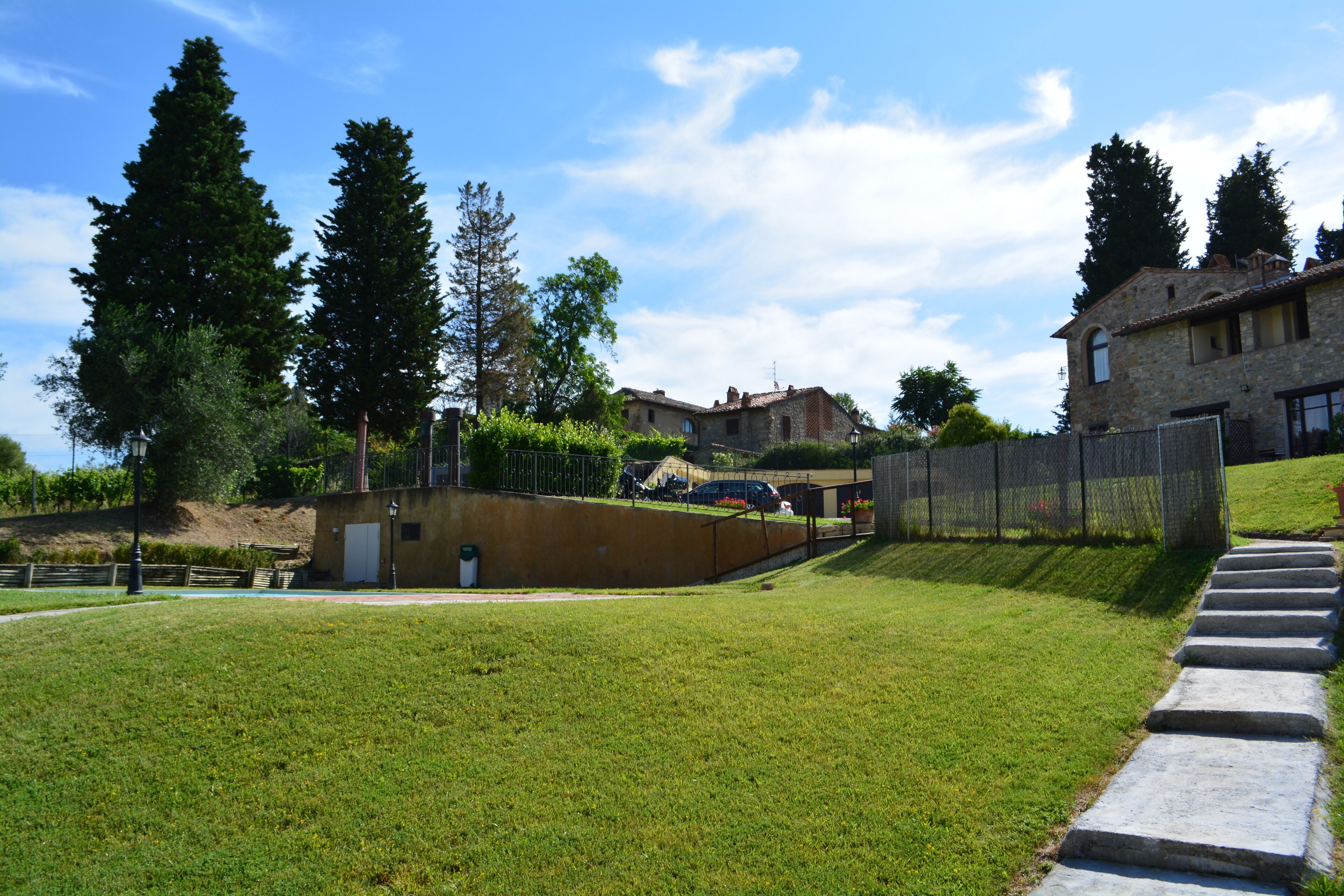 A grassy area with a stone wall, a metal fence, and a stone staircase leading up a hill.