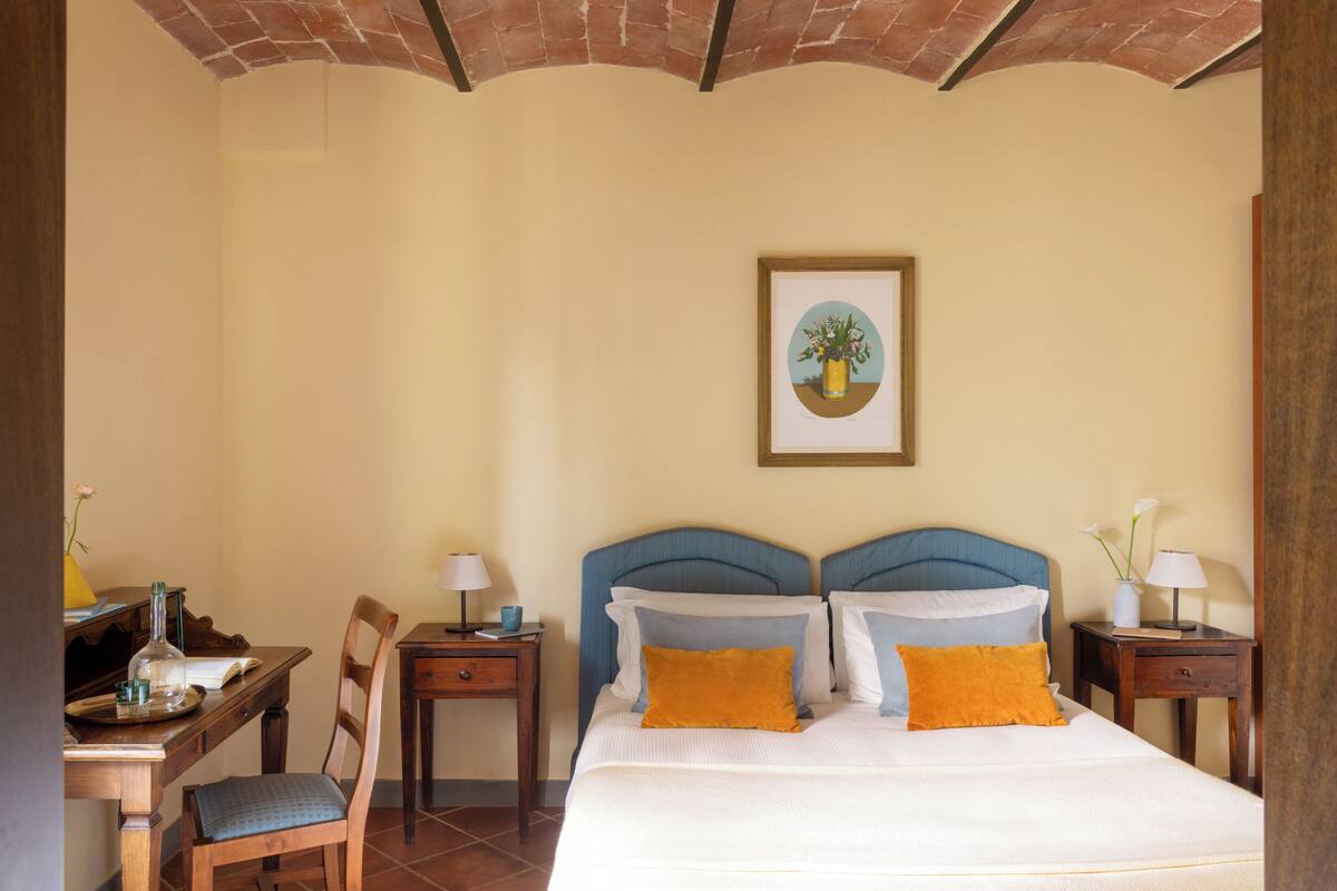 A hotel room with a bed, two bedside tables, a desk with a chair, a framed picture on the wall, and a tiled ceiling.