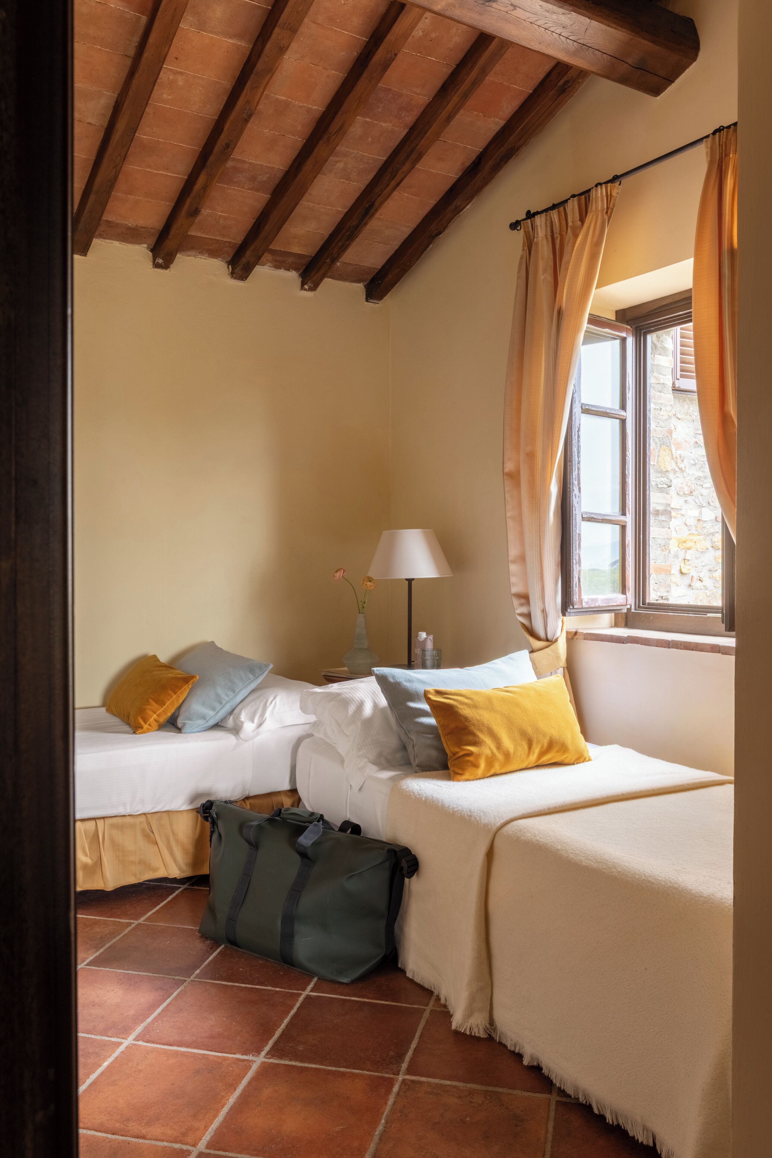 Une chambre avec deux lits, une fenêtre avec des rideaux, une lampe et un sac posé sur le sol.