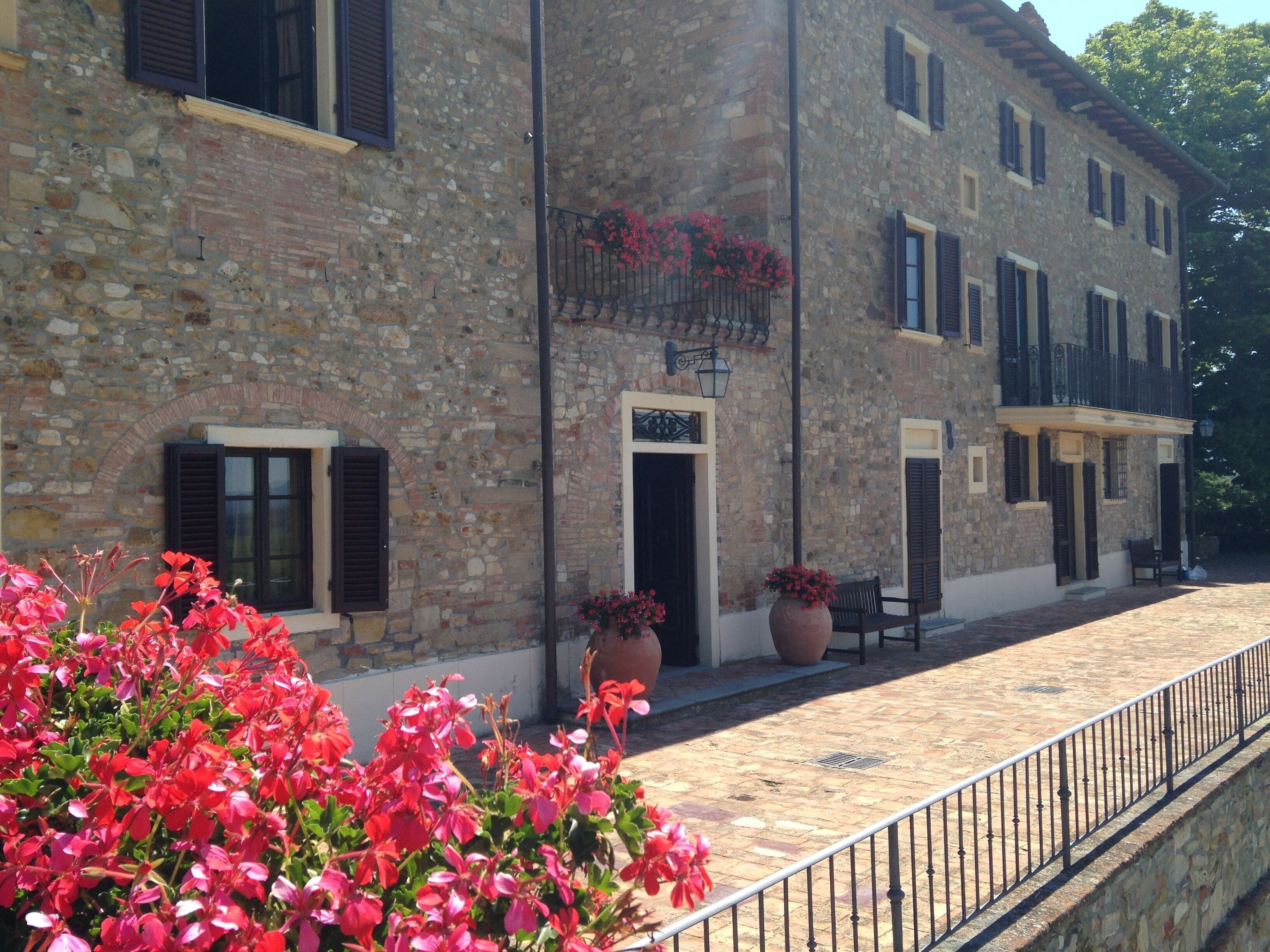A stone building with multiple windows and shutters, a paved courtyard, and potted plants with red flowers.