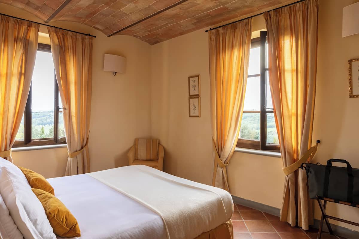 A room with a bed, a chair, a window with curtains, and a bag on a chair.