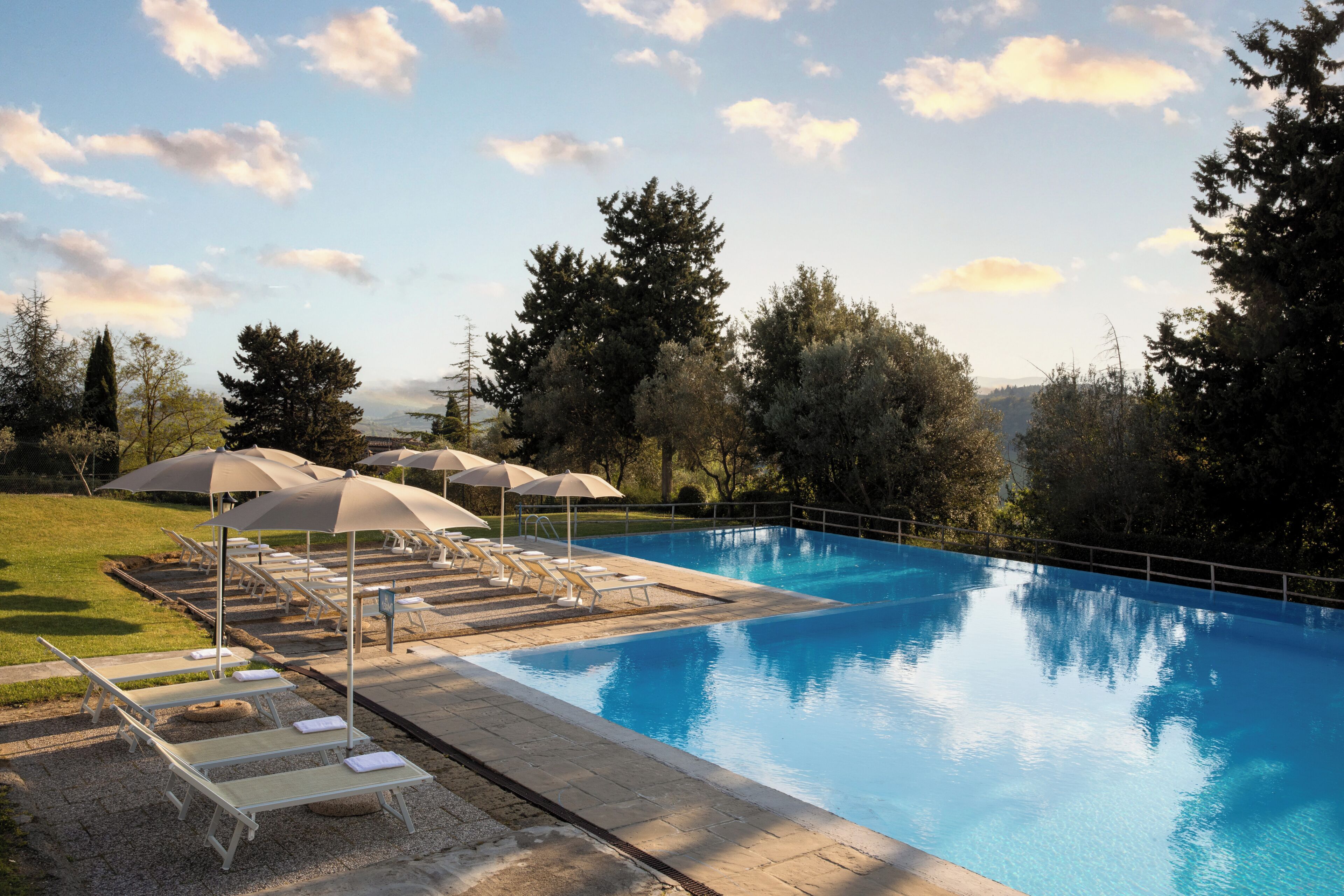 A swimming pool with lounge chairs and umbrellas, surrounded by trees and a clear sky.