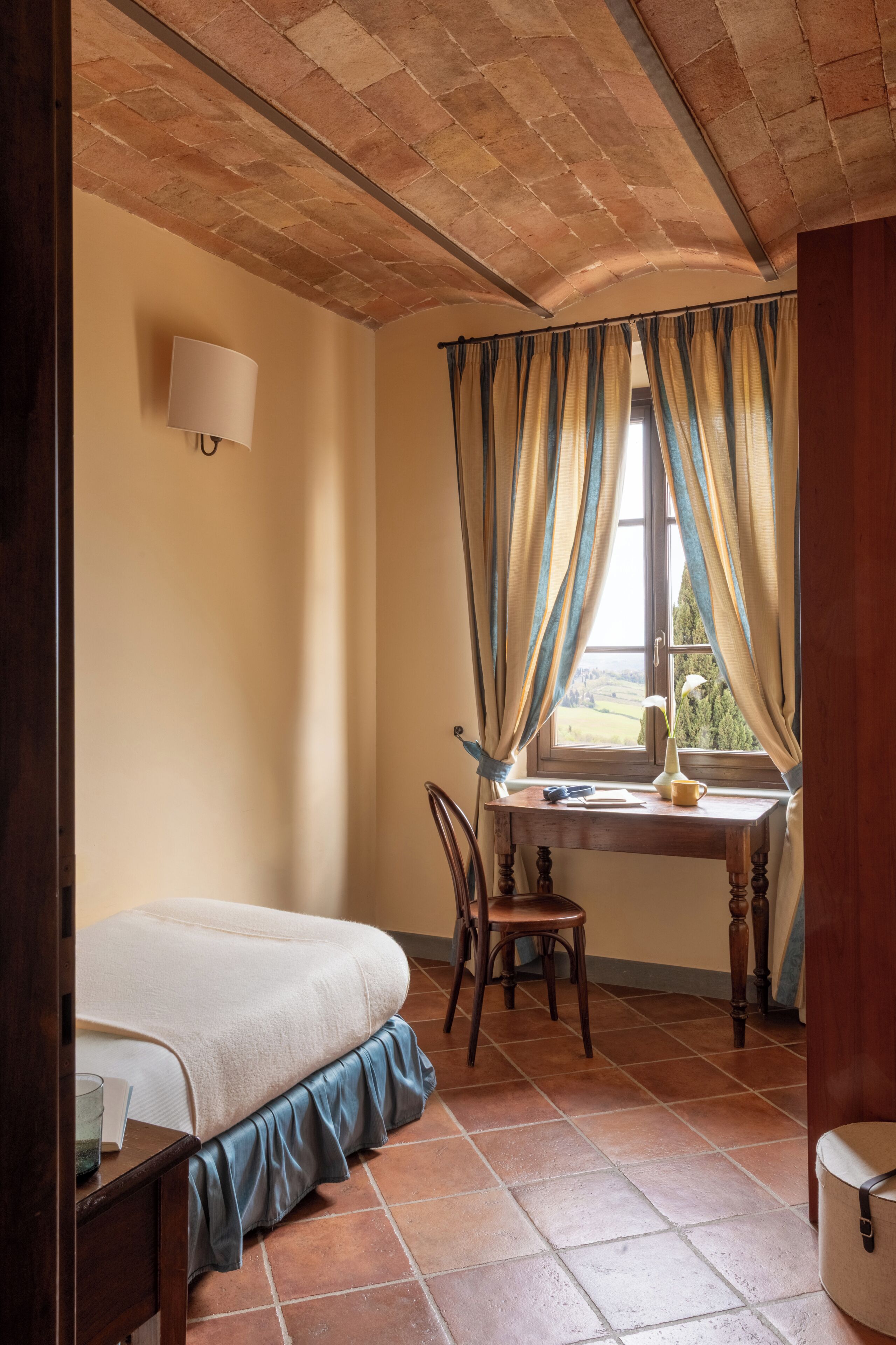 Ein Schlafzimmer mit Bett, Schreibtisch mit Stuhl, einem Fenster mit Vorhängen und einer Wandlampe.