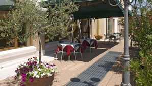 An outdoor seating area with tables and chairs under a green awning, surrounded by potted plants and a metal grate. - Hotel Scaligero (Sommacampagna)
