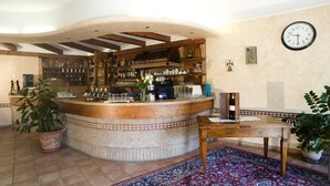 A bar area with a curved wooden counter, shelves with bottles and glasses, a clock on the wall, and a patterned rug. - Hotel Scaligero (Sommacampagna)