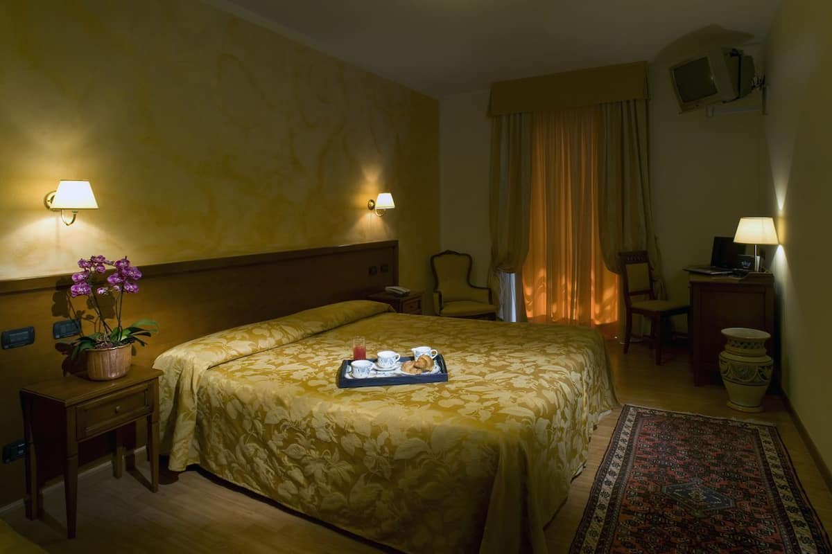 A hotel room with a bed, bedside lamps, a small table with a tray of breakfast, a potted plant, and a rug.