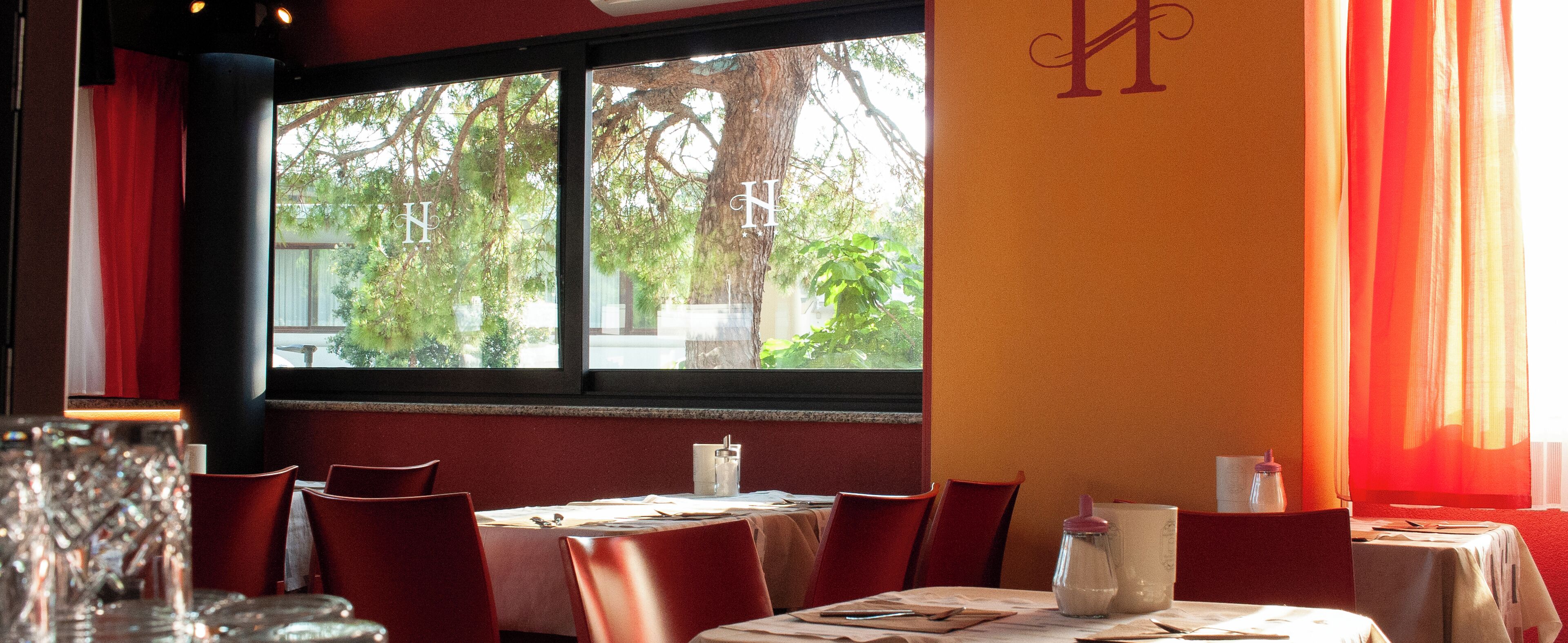 Interior de un restaurante con una mesa preparada para dos, una ventana con vistas a árboles y una pared con una gran letra 'H'.