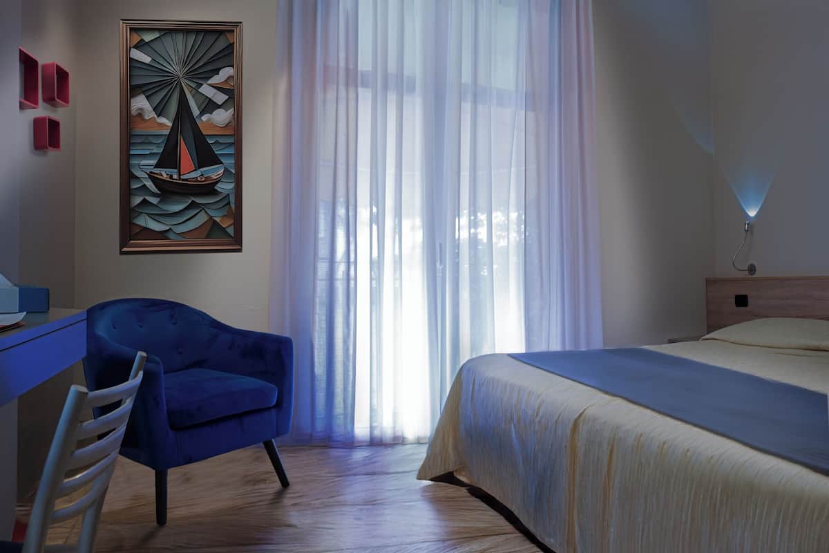 Een hotelkamer met een bed, een bureau met stoel, een schilderij van een zeilboot en een raam met gordijnen.