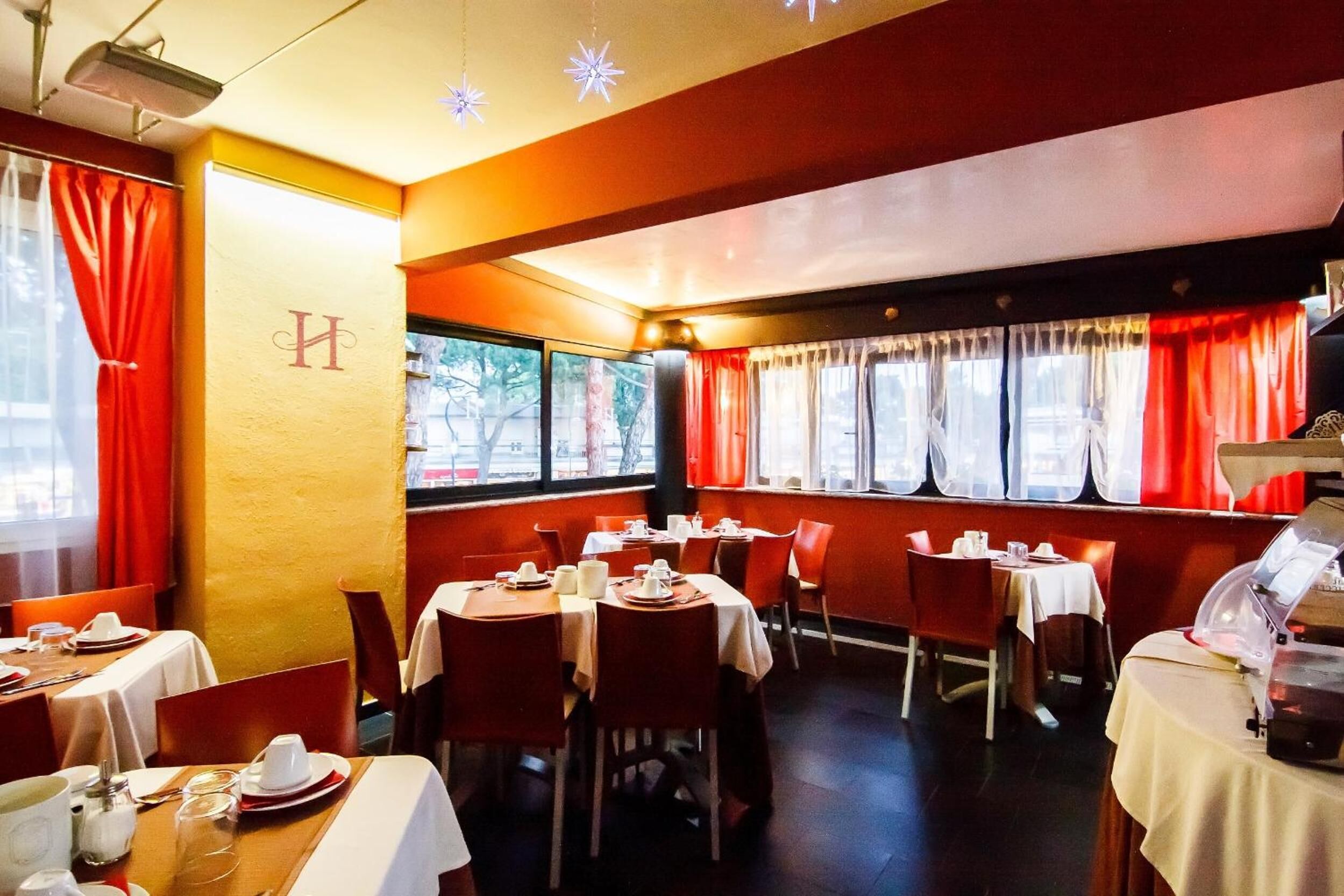 A restaurant with tables set for dining, red curtains, and a 'H' logo on the wall.