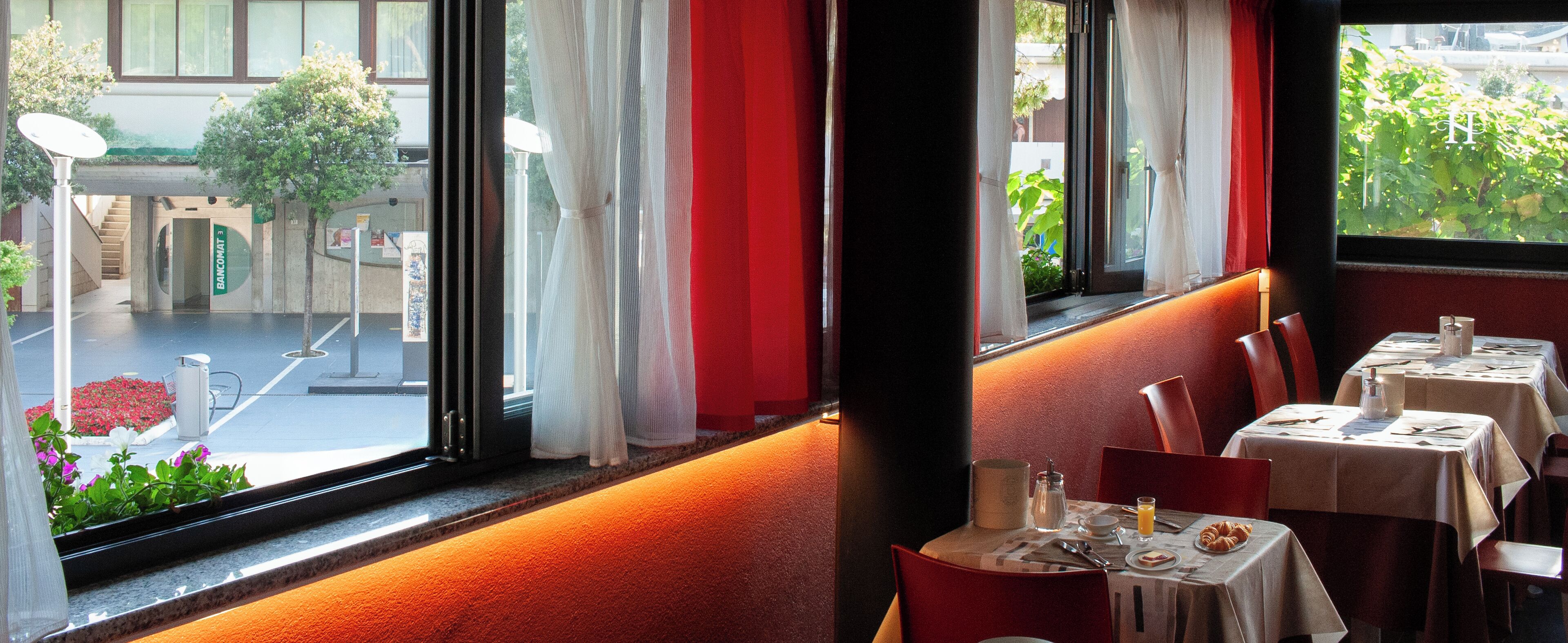 A restaurant with tables set for dining, a view of the street outside, and a building entrance labeled "BANCOMAT".