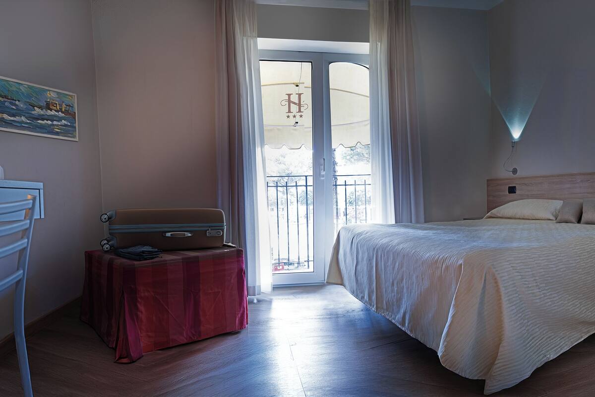 Een slaapkamer met een bed, een koffer, een tafel met een rode tafelkleed, een schilderij aan de muur, en een balkon met gordijnen.
