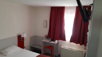 Una habitación de hotel compacta con cama, escritorio, silla, radiador y cortinas rojas.