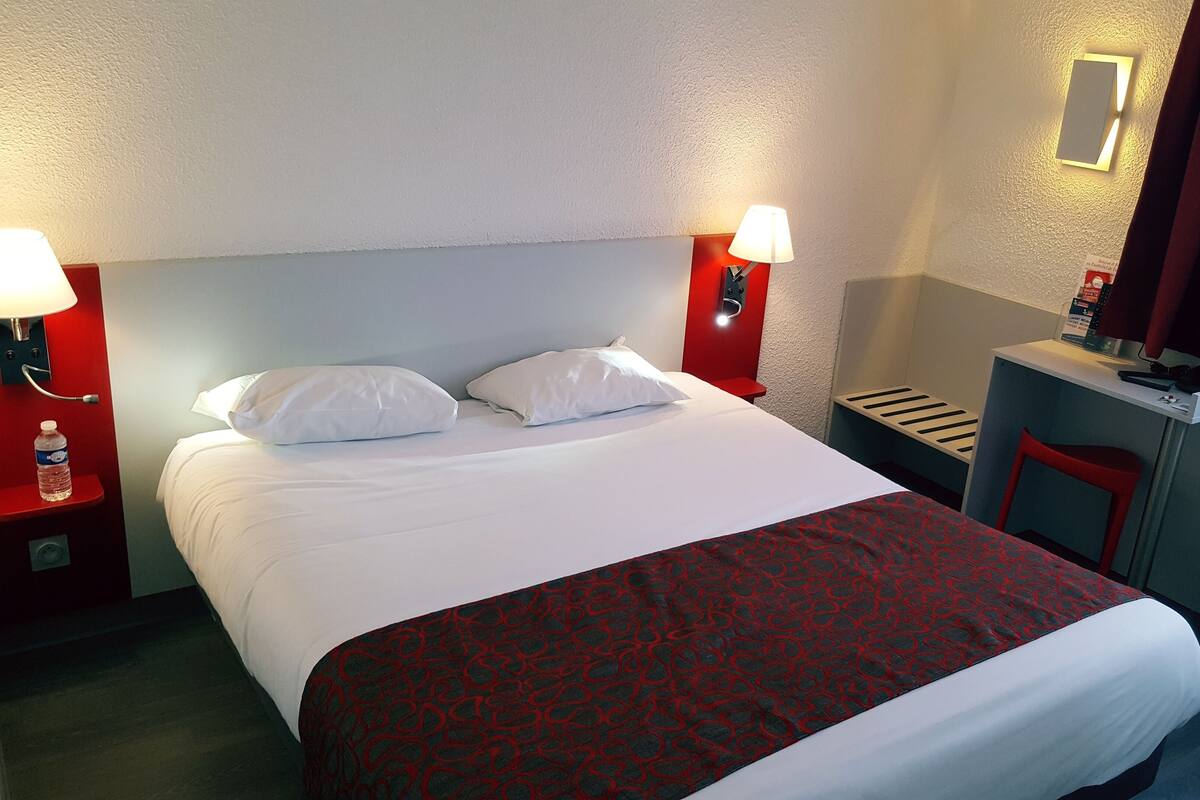 A hotel room with a bed, two bedside lamps, a red headboard, a small desk with a chair, and a red curtain.