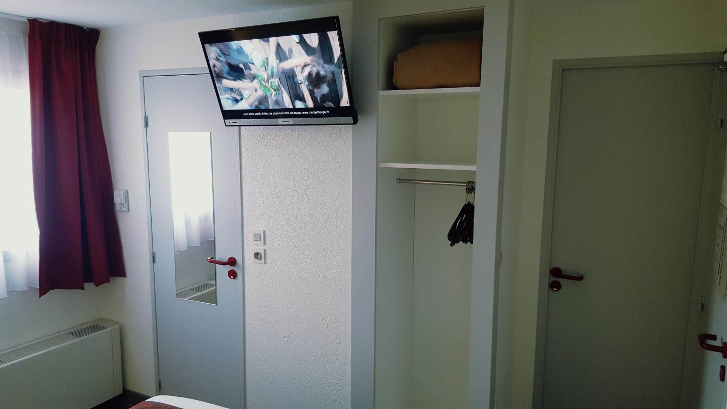 A hotel room with a television mounted on a wall, a door with a red handle, and a window with a red curtain.