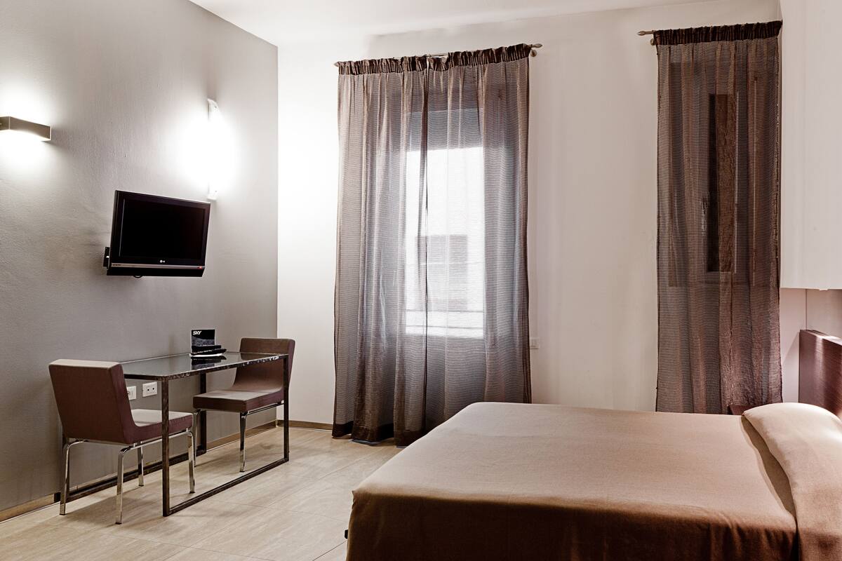Una habitación de hotel con cama, escritorio con silla, televisor montado en la pared y ventana con cortinas.