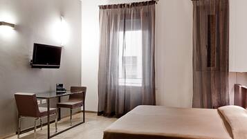Een hotelkamer met een bed, een bureau met stoel, een aan de muur bevestigde televisie en een raam met gordijnen.