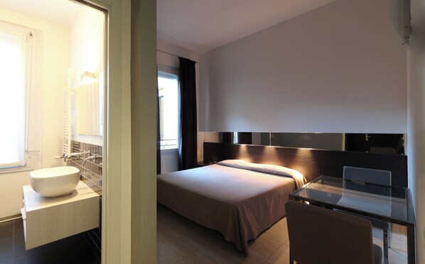 Una habitación de hotel moderna con una cama grande, un escritorio de vidrio y un baño con lavamanos y espejo blancos.