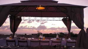 An outdoor dining area with tables set for two, under a pergola with a view of the sunset and distant landscape. - A Giummara (Marsala)