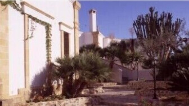 A courtyard with stone steps leading to a building with a white facade and a chimney. There are plants and shrubs around the area. - A Giummara (Marsala)