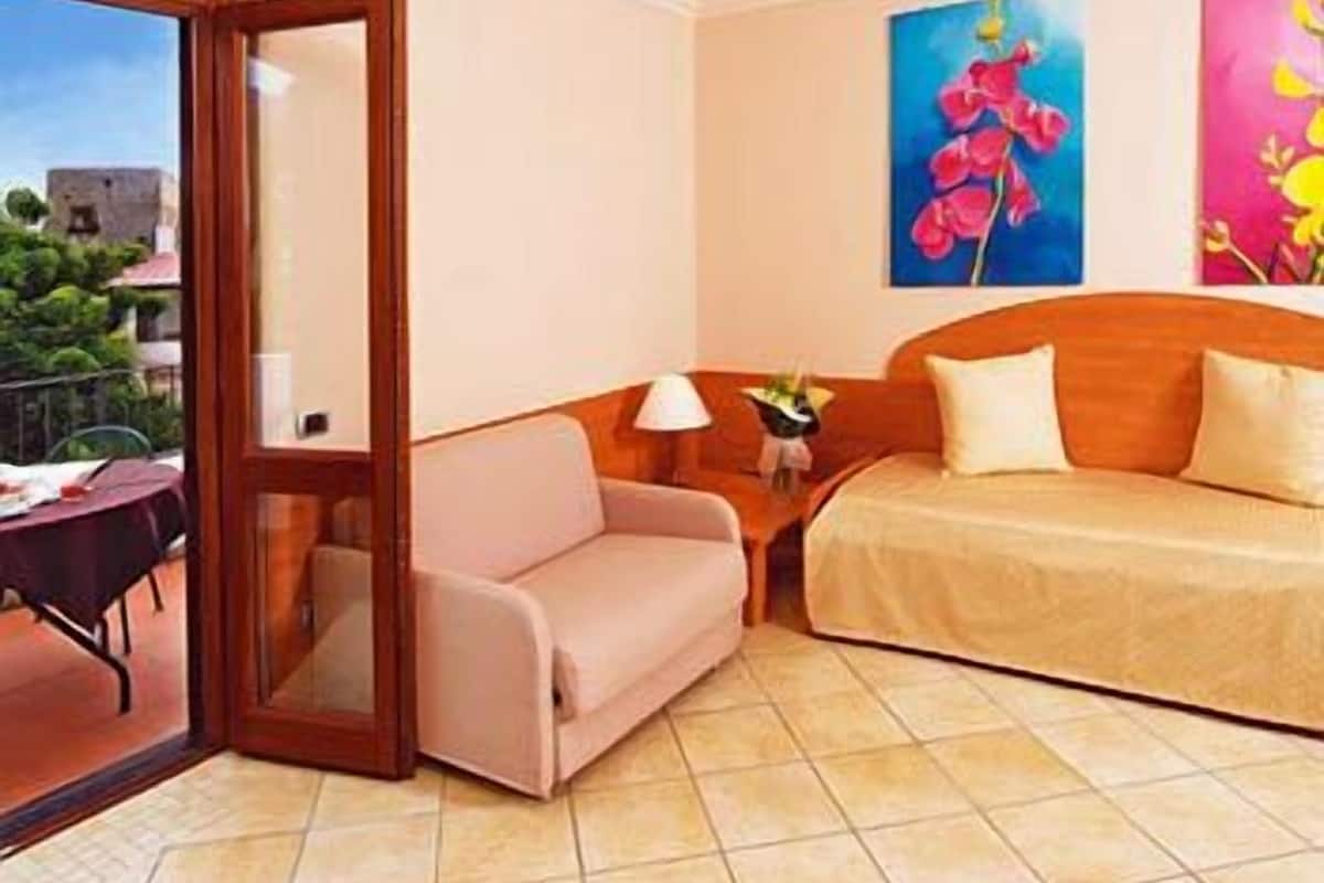 A hotel room with a bed, a sofa, a table with a lamp, and artwork on the walls.