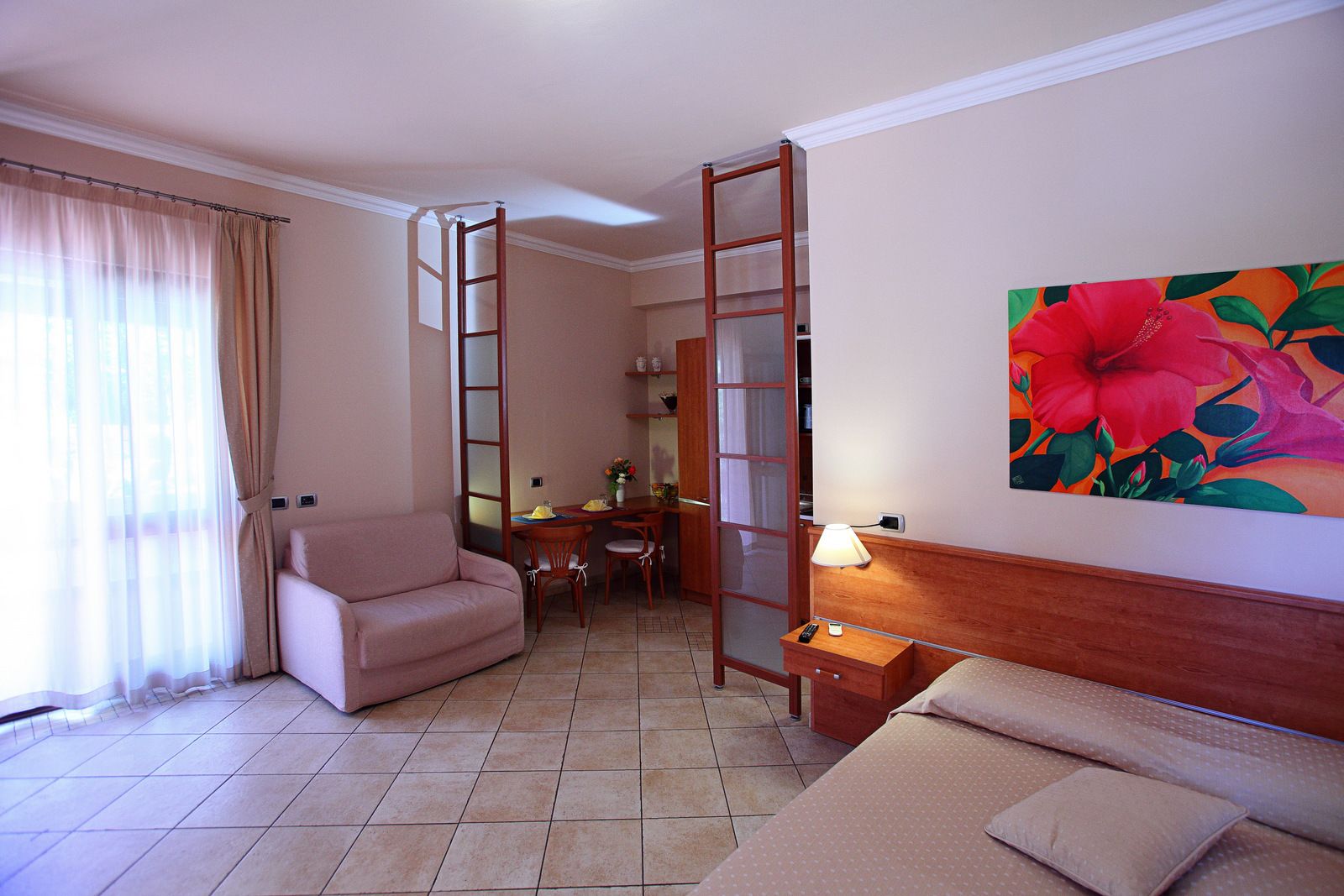 A hotel room with a sofa, a desk with chairs, a ladder, a bed, and a painting of a flower on the wall.