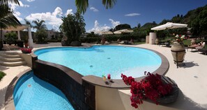 Ein Pool mit geschwungener Kante, umgeben von Liegestühlen und Sonnenschirmen, sowie eine Steinmauer mit einem kleinen Pool.
