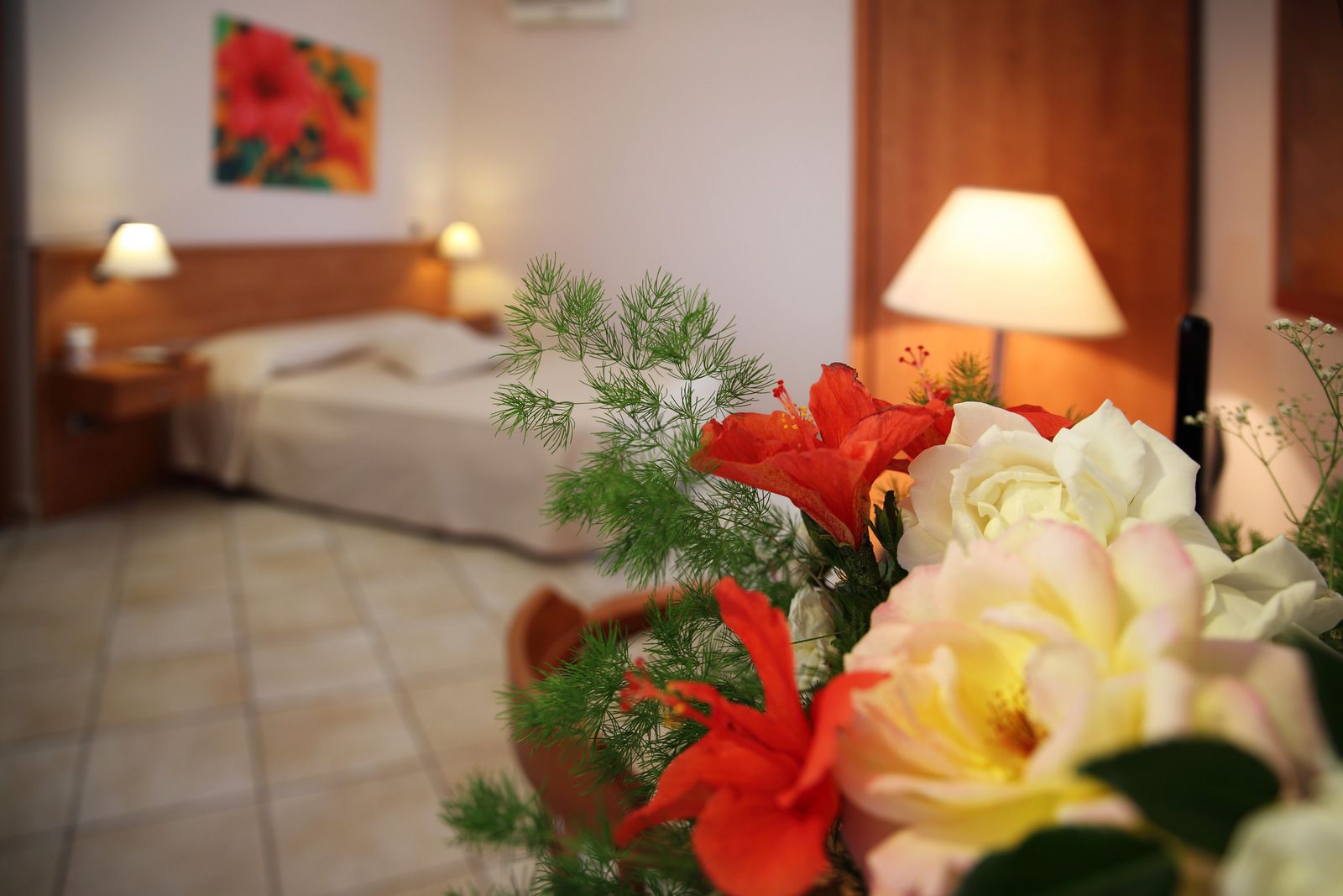A hotel room with a bed, bedside lamps, a floral arrangement, and a painting on the wall.