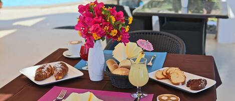 Una mesa servida con cruasanes, pan, una bebida y una taza de café, con una piscina al fondo.