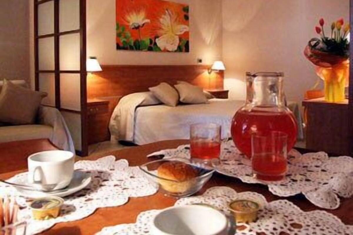 A hotel room with a neatly made bed, a painting on the wall, a bedside table with a lamp, and a table set with breakfast items.