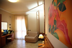 Habitación de hotel con un sillón color beige, un escritorio con computadora, una lámpara y un cuadro grande con motivos florales en la pared.