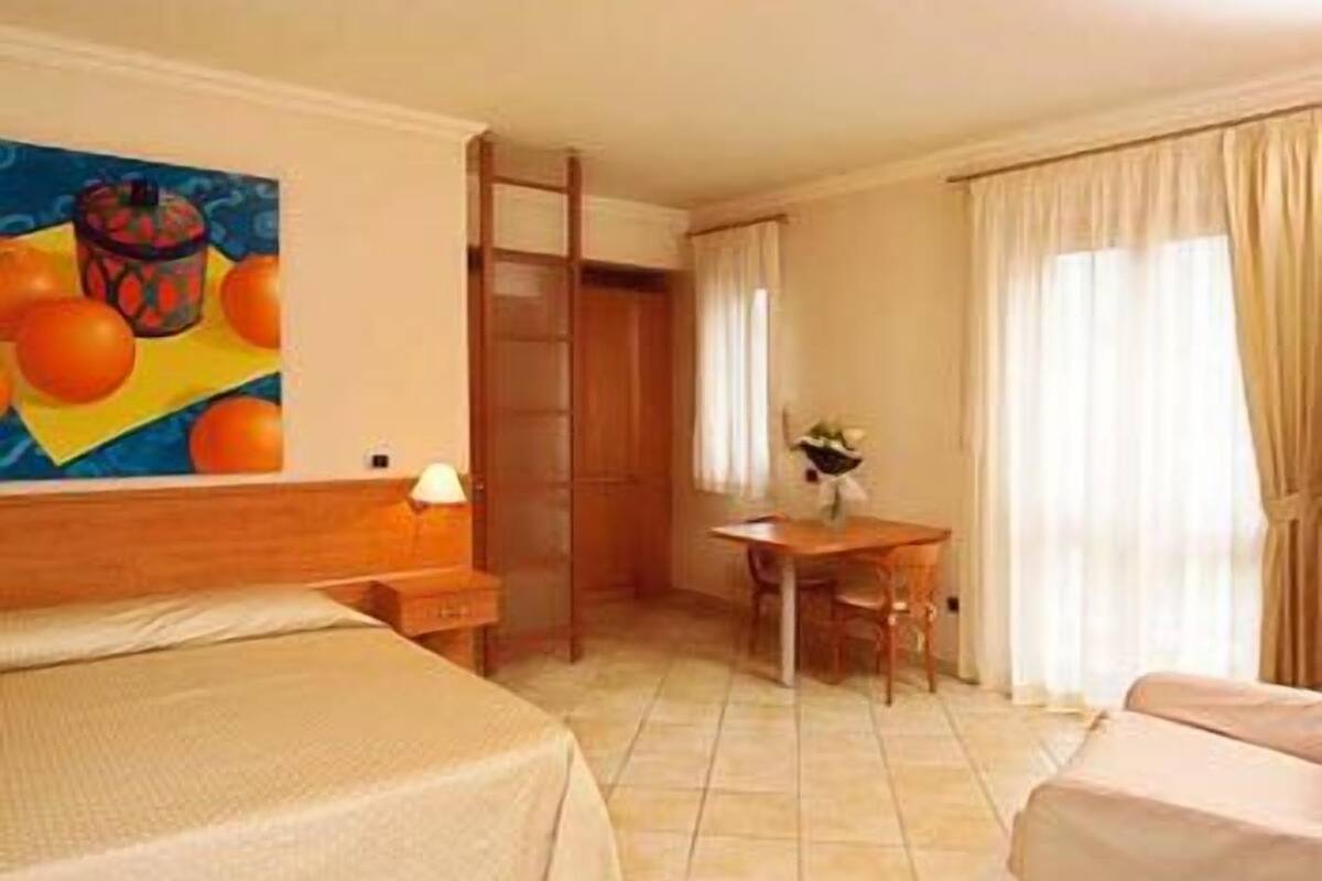 A hotel room with a bed, a desk, a chair, a wardrobe, and a painting on the wall.