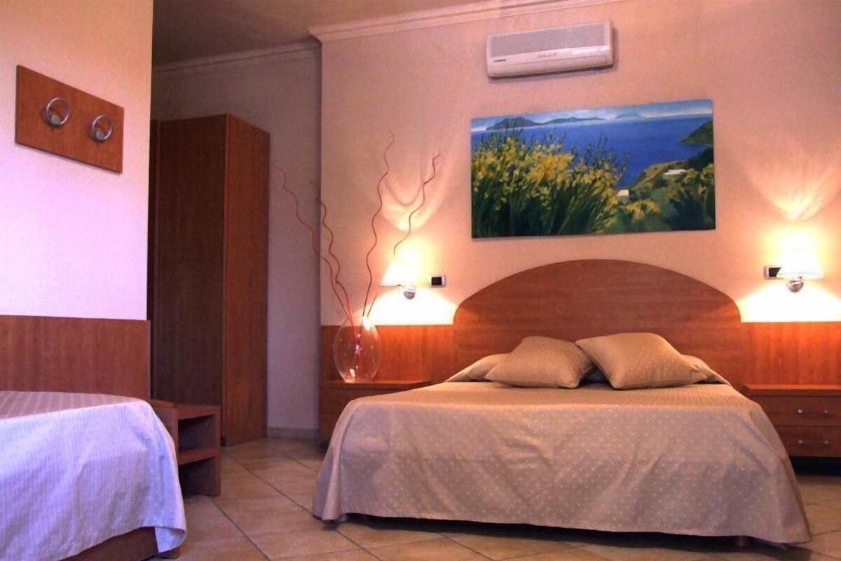 A hotel room with a large bed, a smaller bed, a painting of a landscape, and a wall-mounted air conditioner.