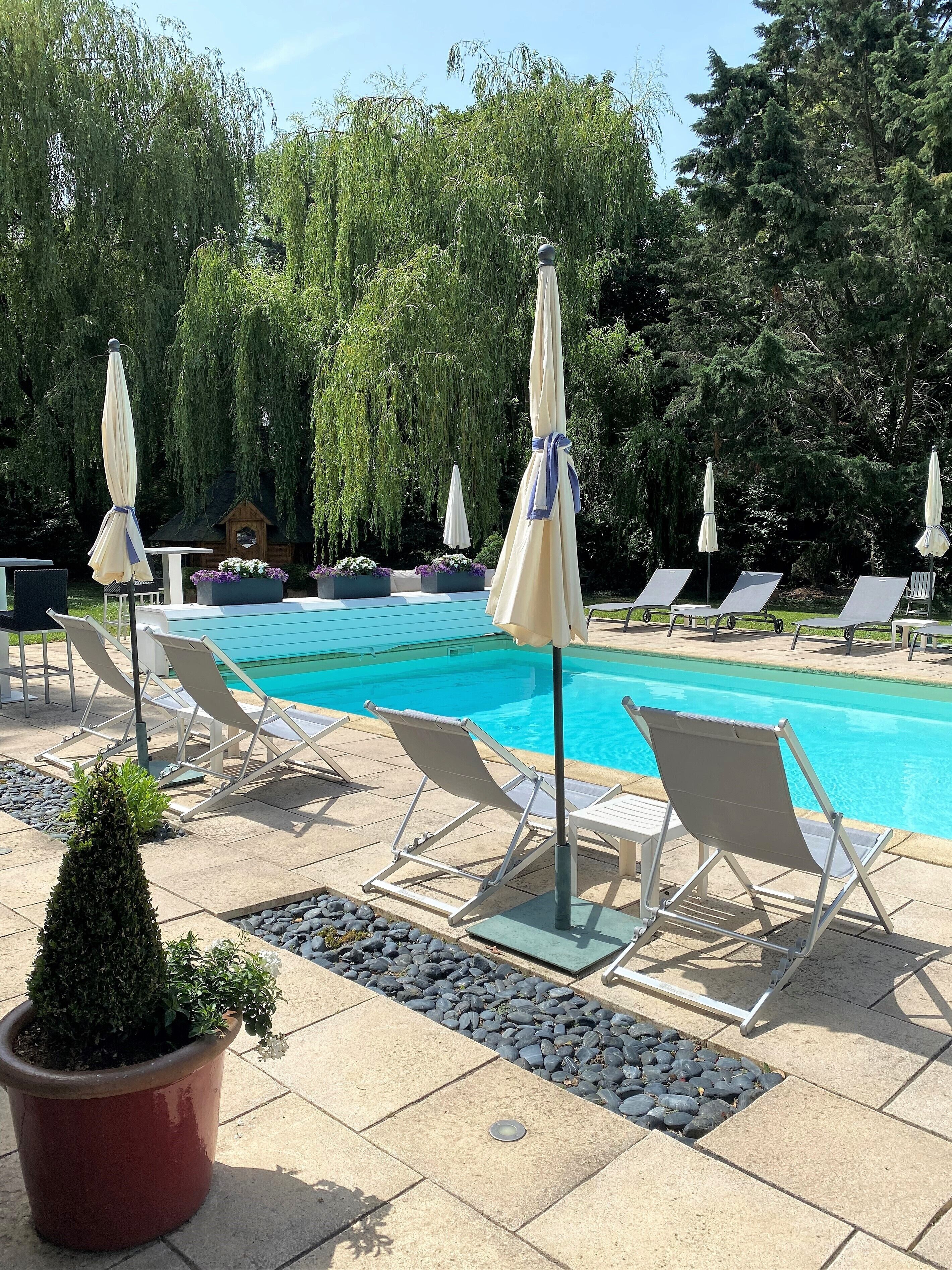 Zona junto a la piscina con sillones y sombrillas, rodeada de árboles y una estructura de madera.