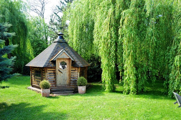 A small wooden cabin with a sloped roof, surrounded by lush greenery and a weeping willow tree. - Les Jardins d'Adalric (Obernai)