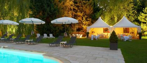 Un área junto a la piscina con sillones de descanso y sombrillas, un comedor cubierto por una carpa y un césped bien cuidado.