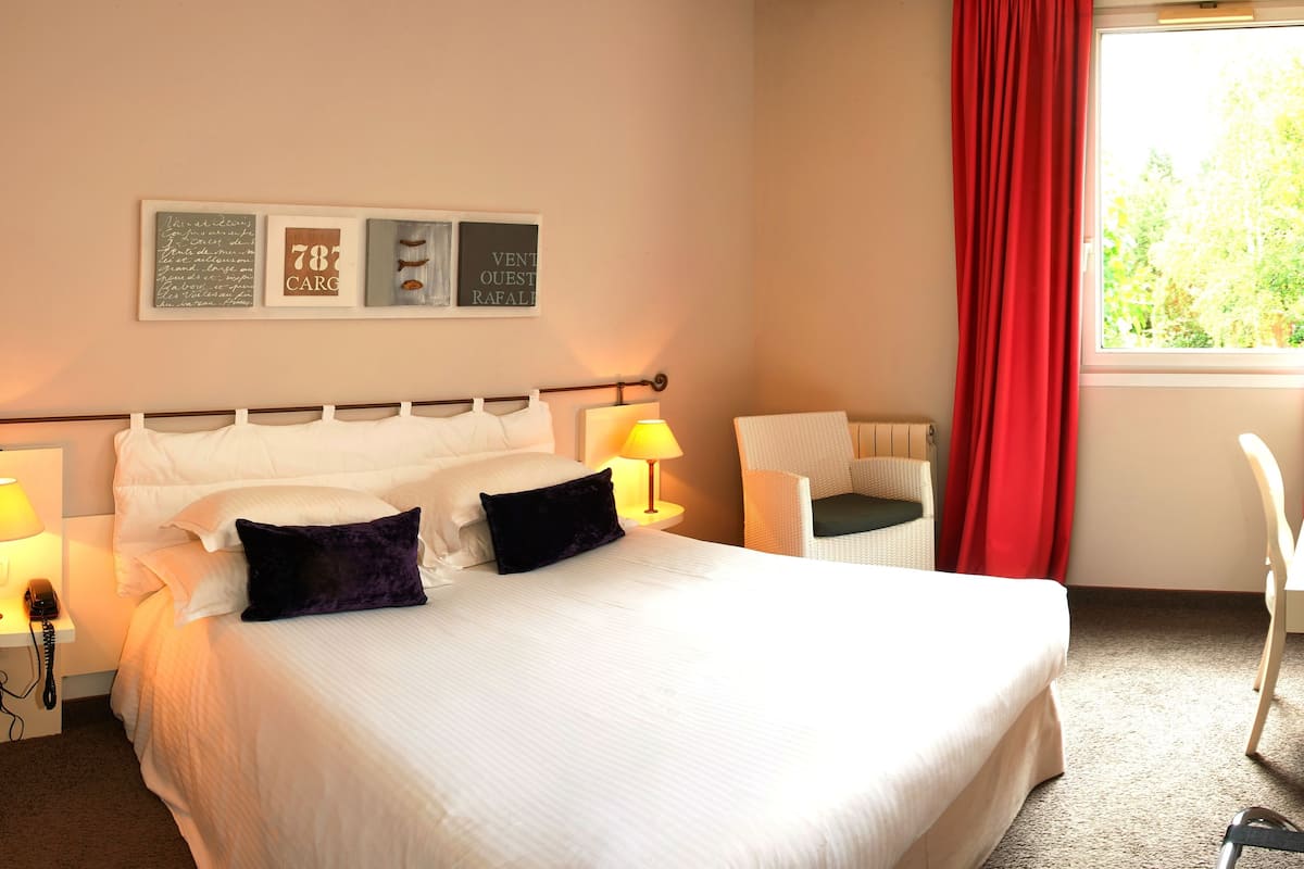 A hotel room with a bed, two bedside lamps, a desk with a chair, a TV, and a window with red curtains.