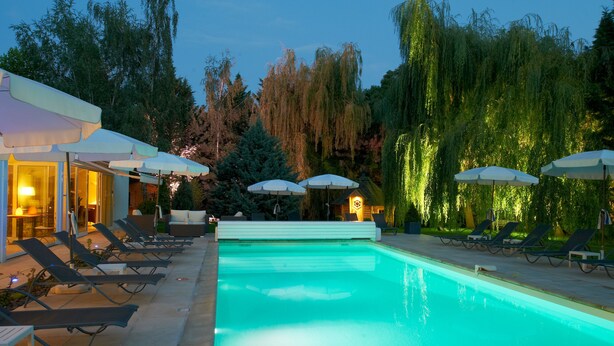 A swimming pool with lounge chairs and umbrellas, surrounded by trees and a building with lights.