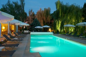A swimming pool with lounge chairs and umbrellas, surrounded by trees and a building with lights. - Les Jardins d'Adalric (Obernai)