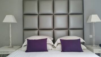 A neatly made bed with two white pillows and two purple accent pillows, flanked by two white bedside tables with lamps.