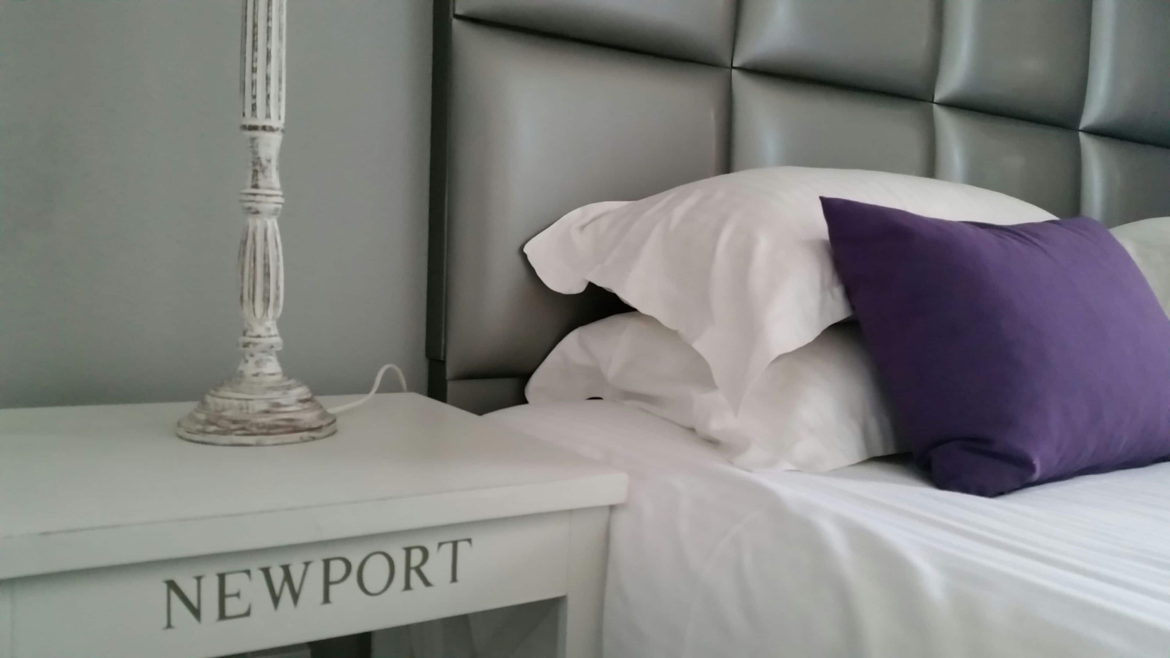Una cama bien hecha con almohadas blancas y moradas, una mesita de noche blanca con el nombre 'NEWPORT' grabado, y un cabecero moderno.