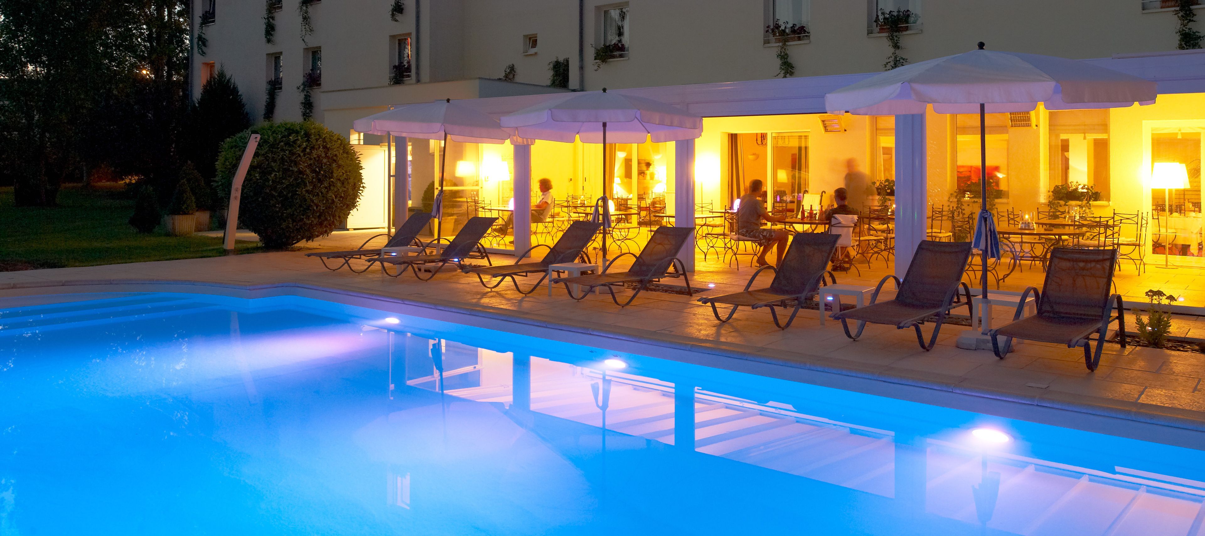 Una piscina con sillas de descanso y sombrillas, un restaurante con mesas y sillas, y un edificio con ventanas y plantas.