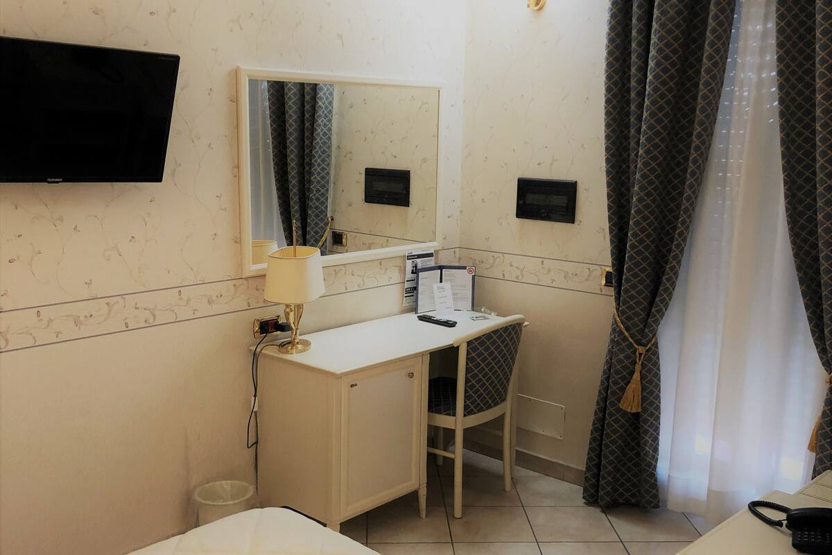 A hotel room with a bed, a desk with a chair, a television mounted on the wall, a mirror, and a window with curtains.