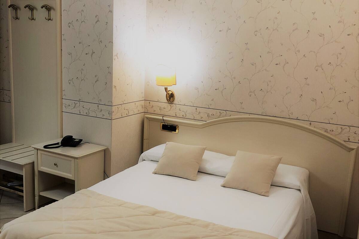 A neatly made bed with a beige comforter and pillows, a bedside table with a lamp, and a mirror on the wall.