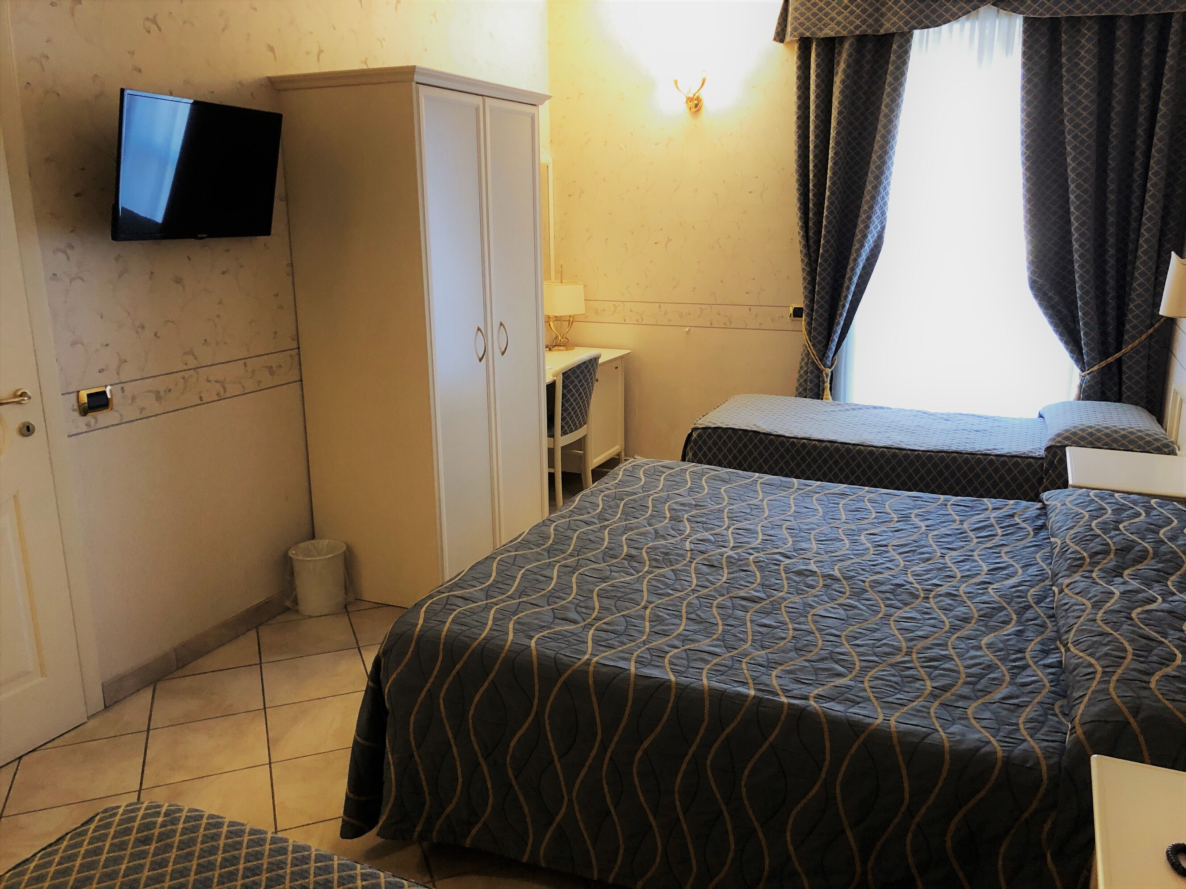 A hotel room with two beds, a TV mounted on the wall, a wardrobe, and a small desk with a lamp.