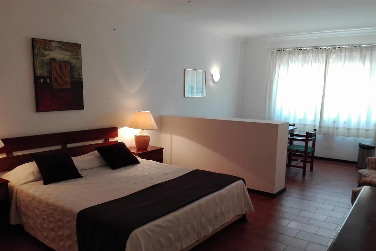 A hotel room with a bed, a nightstand with a lamp, a painting on the wall, a window with curtains, and a small table with chairs.