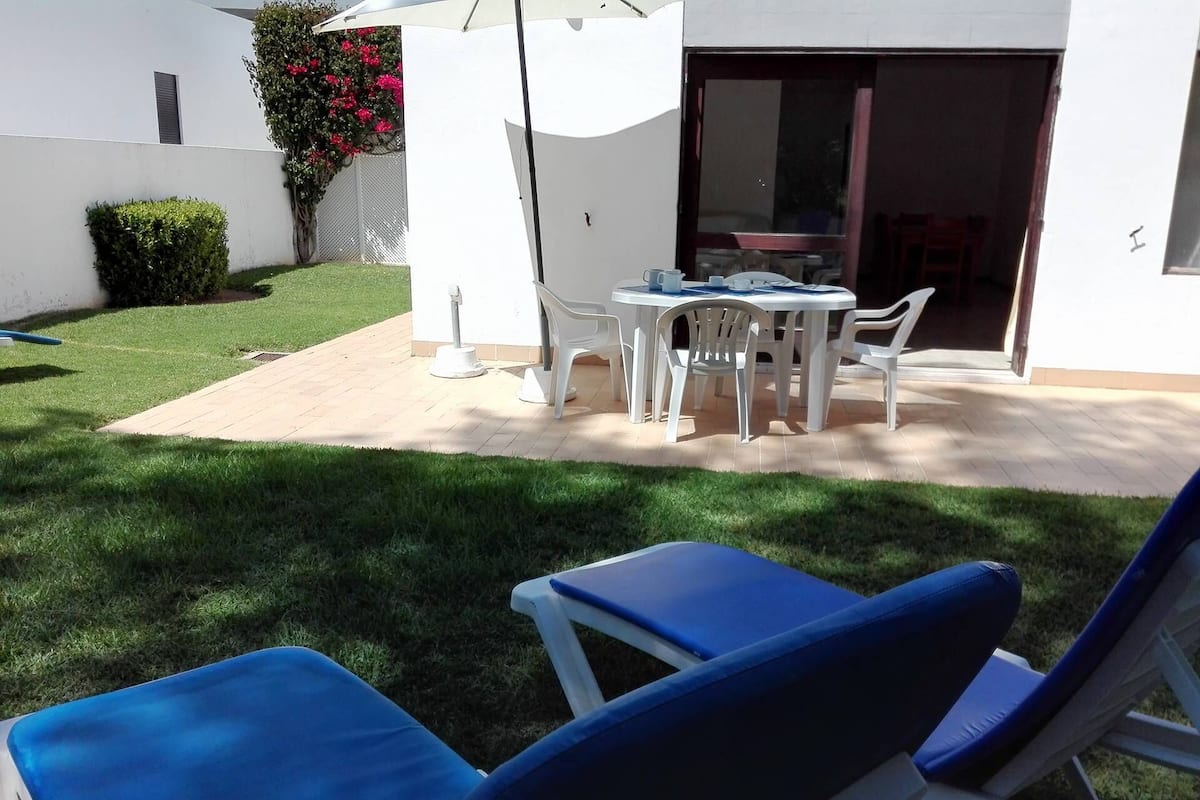 A patio area with a table and chairs, a small garden, and a white building with a sliding glass door.