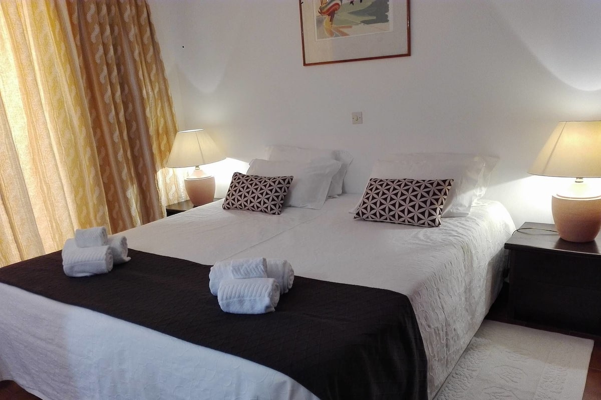 A neatly made bed with white linens and two pillows, a black bedspread, and two towels on the bed. There are two bedside tables with lamps, a wall clock, and a framed picture above the bed.