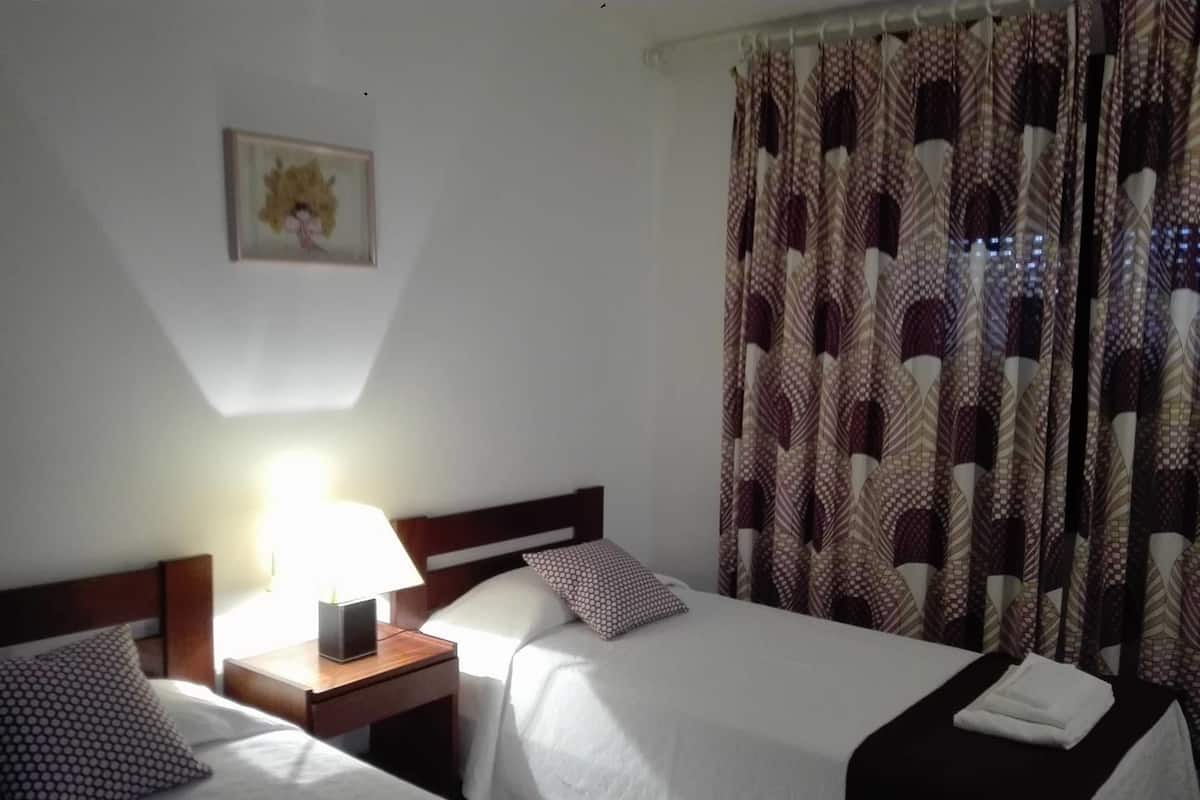 A hotel room with two beds, a nightstand with a lamp, patterned curtains, and a framed picture on the wall.