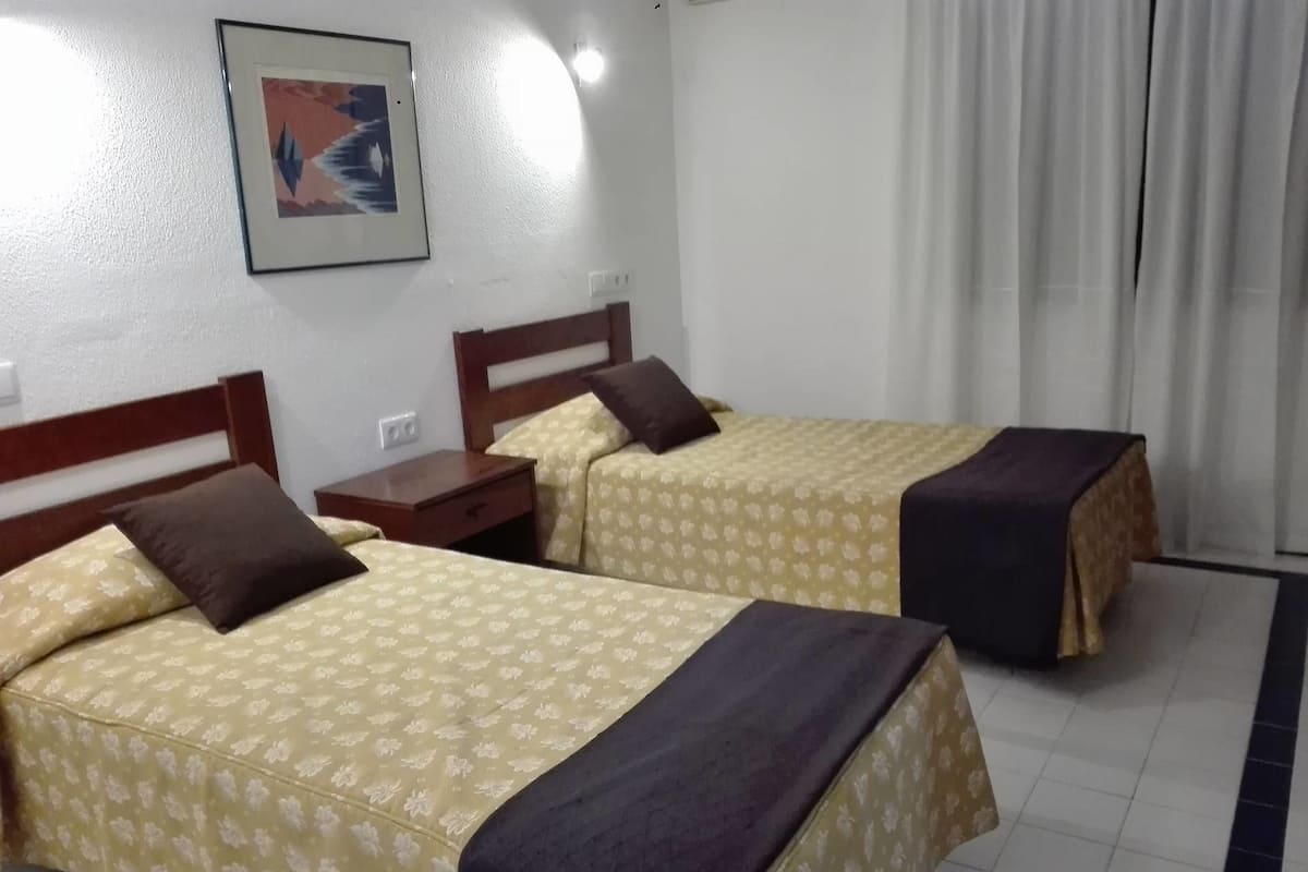A hotel room with two beds, each with a floral bedspread and a dark throw blanket. There is a nightstand with a lamp between the beds, and a framed picture on the wall.
